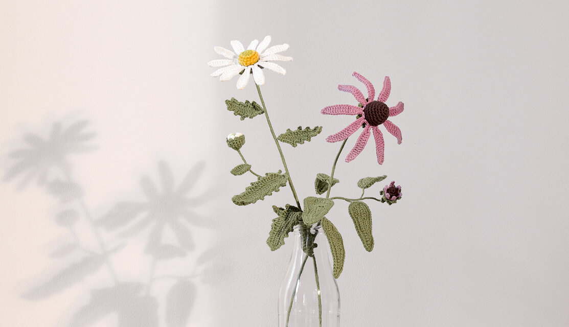 Material Ricorumi In the Meadow Wiesenmargerite, gehäkelte Blumen in Glasvase, filigrane Blüten und Blätter als dekoratives DIY-Projekt vor hellgrauem Hintergrund