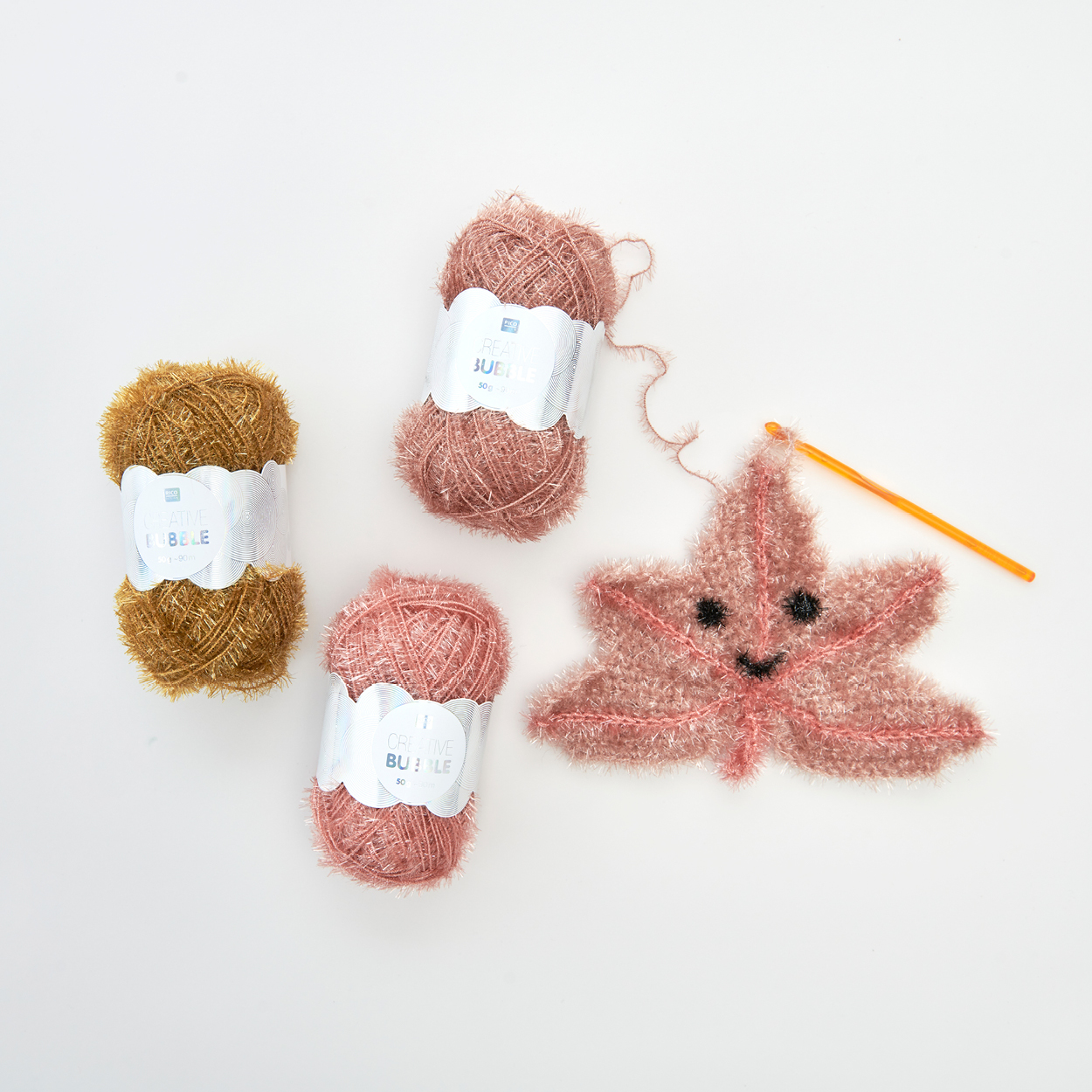 Ein gehäkeltes Blatt aus Schwammgarn mit Häkelnadel und drei Schwammgarnknäuel. Flatlay von Produkten.