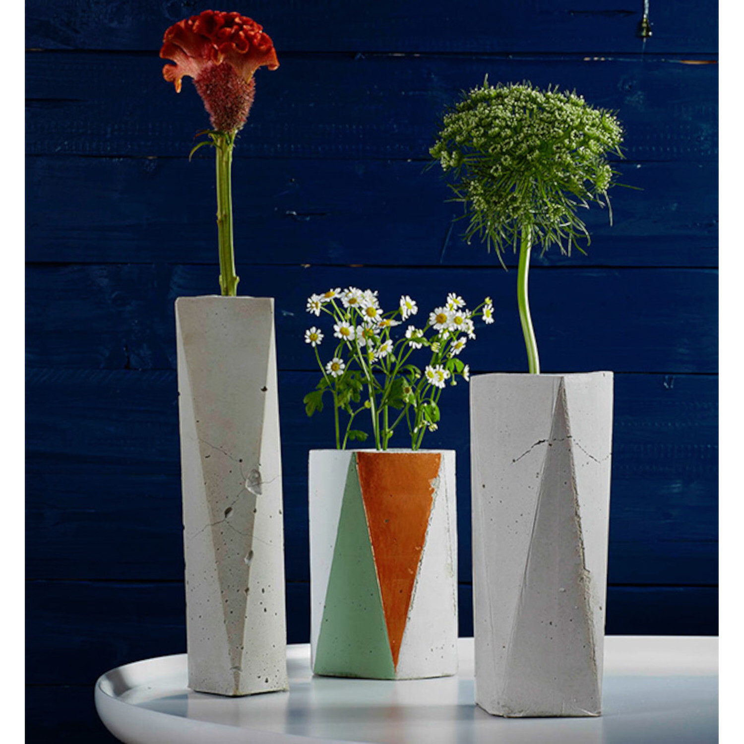 Beton - Techniken und Ideen für Einsteiger und Fortgeschrittene: Drei moderne Betonvasen mit geometrischen Mustern auf weißem Tisch vor dunkelblauem Hintergrund.
