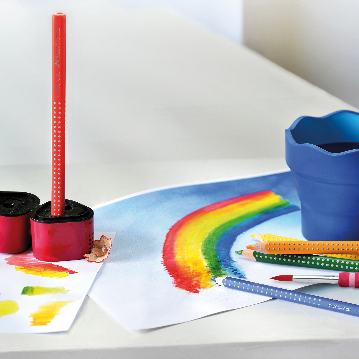 Jumbo Grip Farbstift von Faber Castell in Mauve liegt neben buntem Regenbogenbild und Malzubehör.