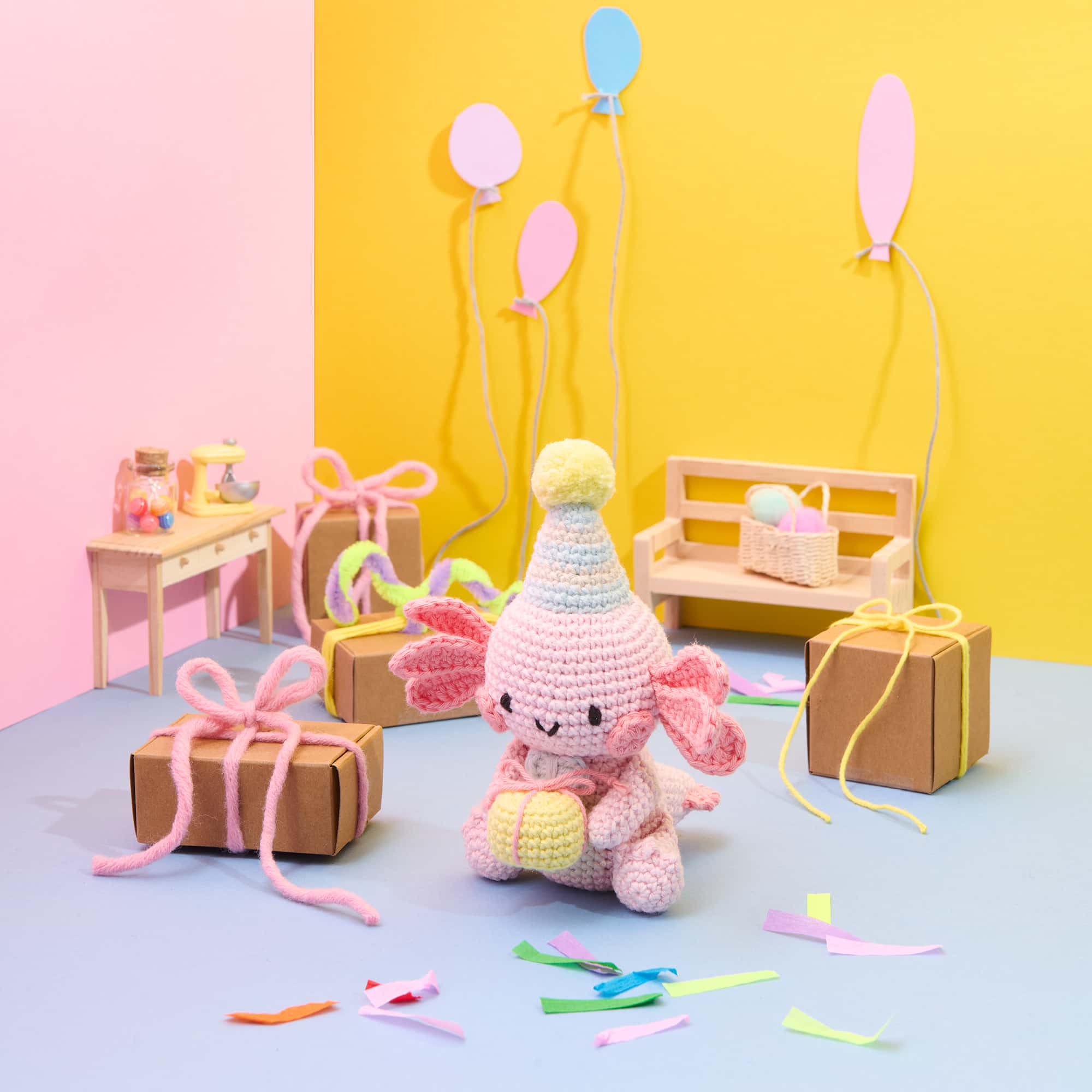 Ein gehäkeltes Axolot mit Partyhut hält ein Geschenk und sitzt vor einer bunt dekorierten Szene mit Partydekoration.