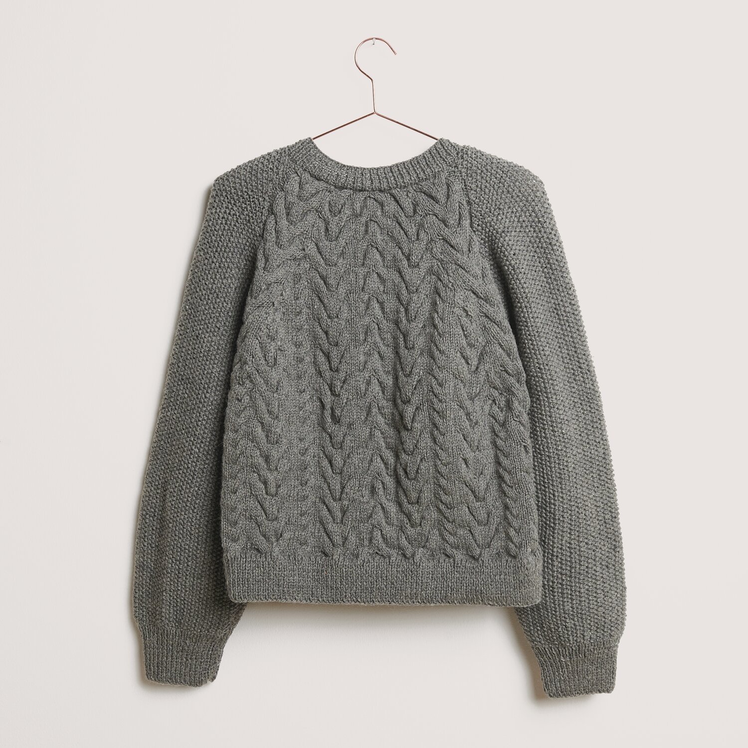 Die Neue Masche No. 14 Winter von idee. Creativmarkt Essentials Soft Merino aran Pullover in Mittelgrau, gestrickt aus Rico Design Wolle, hängt an einem Kleiderbügel vor neutralem Hintergrund.