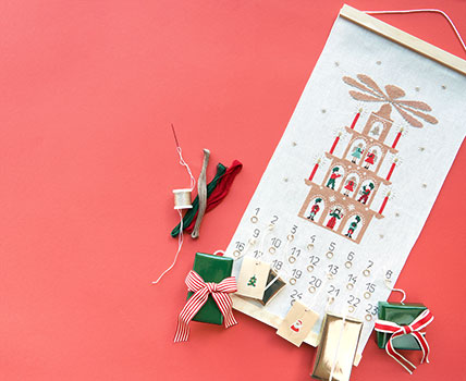 Ein Adventskalender mit einer Lichterpyramide und Zahlen bestickt, sowie kleinen Ringen an welchem Geschenke hängen. Flatlay von Produkten.