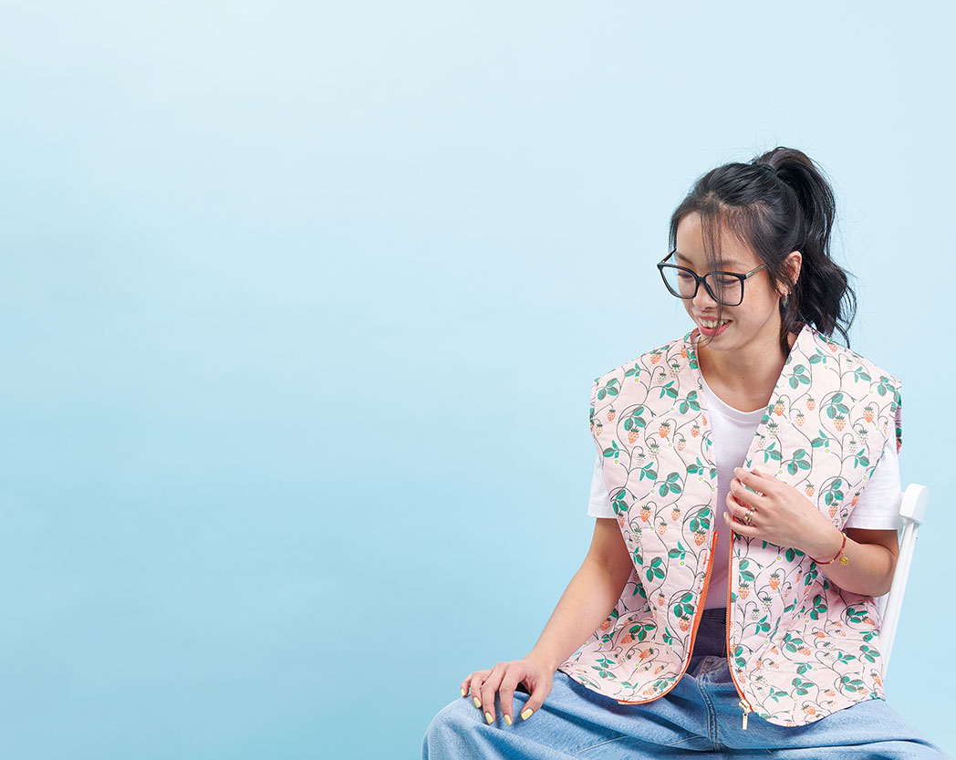 Eine Frau sitzt und trägt eine Weste aus einem pinken und grünen Stoff mit Motiv. Fotosetting blaue Wand.