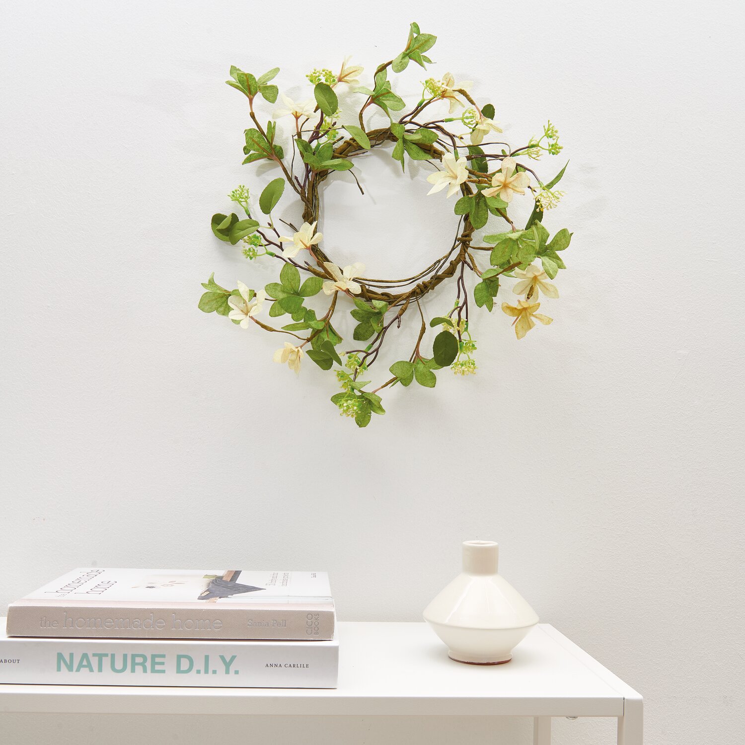 Kranz mit Blüten creme-weiß 40cm, dekorativ an weißer Wand, umgeben von grünen Blättern.