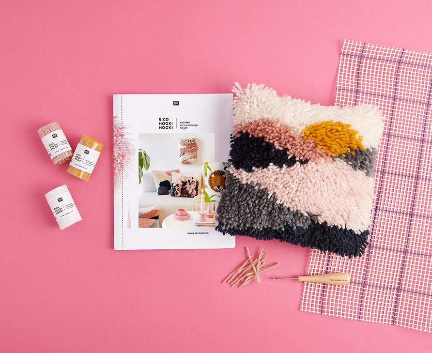 Ein geknüpftes Kissen, ein Knüpf-Heft, verschiedenes Knüpfgarn, Knüpfstramin und ein Knüpfhaken. Flatlay von Produkten.