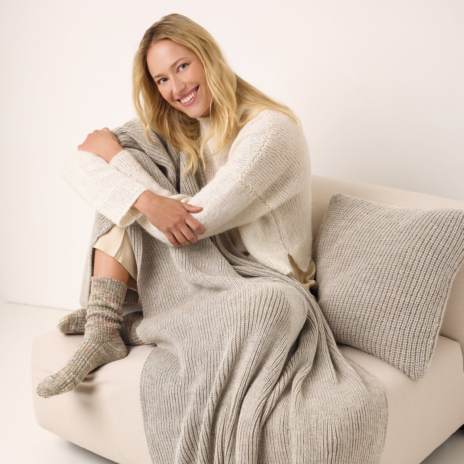 Strickset Socken Modell 04 aus Die Neue Masche No. 13 von idee. Creativmarkt Strickset Socken Modell 04: Frau in beigem Strickoutfit sitzt auf Sofa, trägt gestrickte Socken.