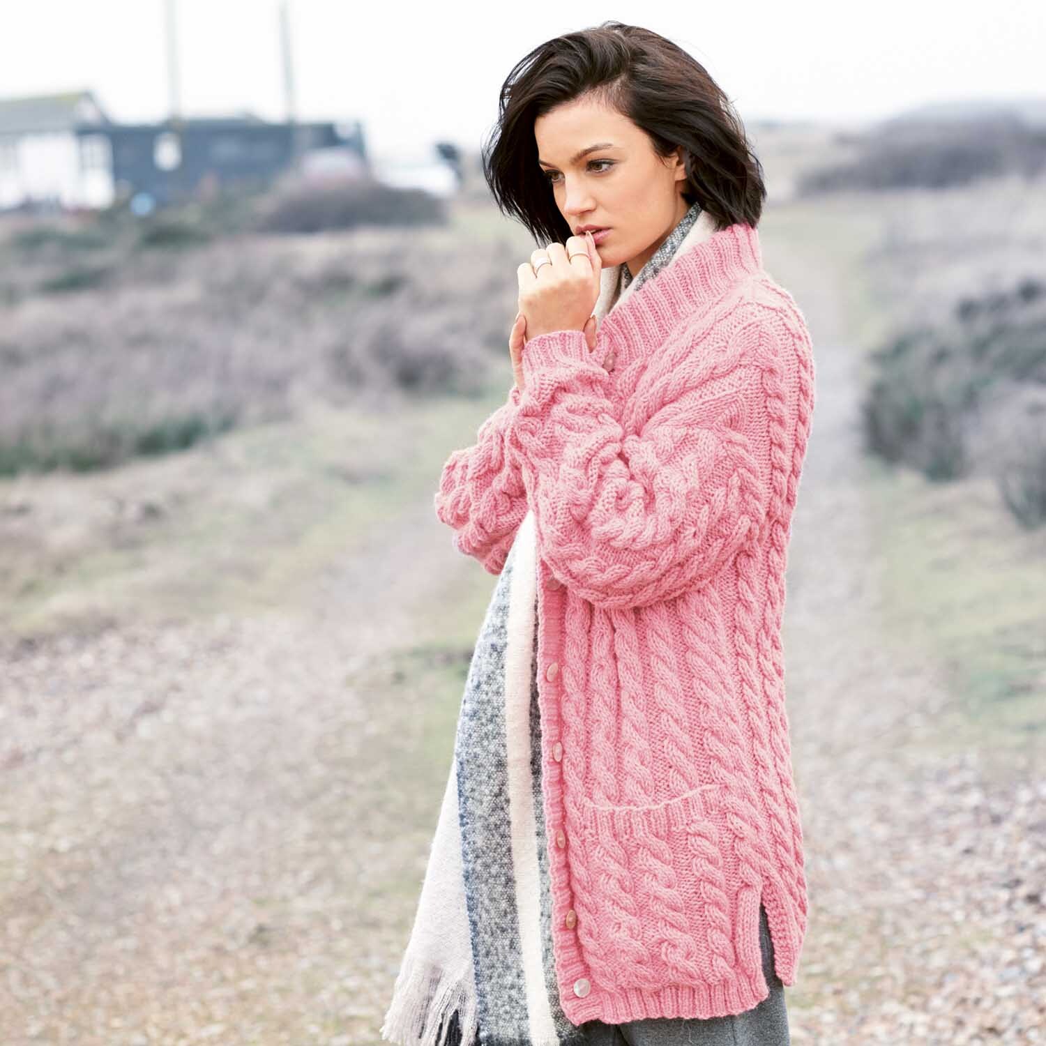 Strickanleitung Zopfmuster-Jacke: Frau in rosa Zopfmuster-Jacke aus Alpaka-Wollmischung in natürlicher Umgebung.