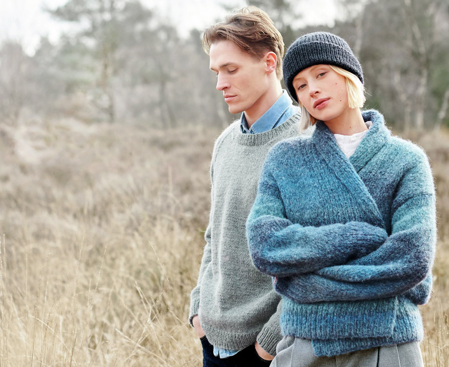 Frau trägt blaue Strickjacke und -mütze, Mann trägt grauen Strickpullover - sie stehen vor einer Waldlichtung.