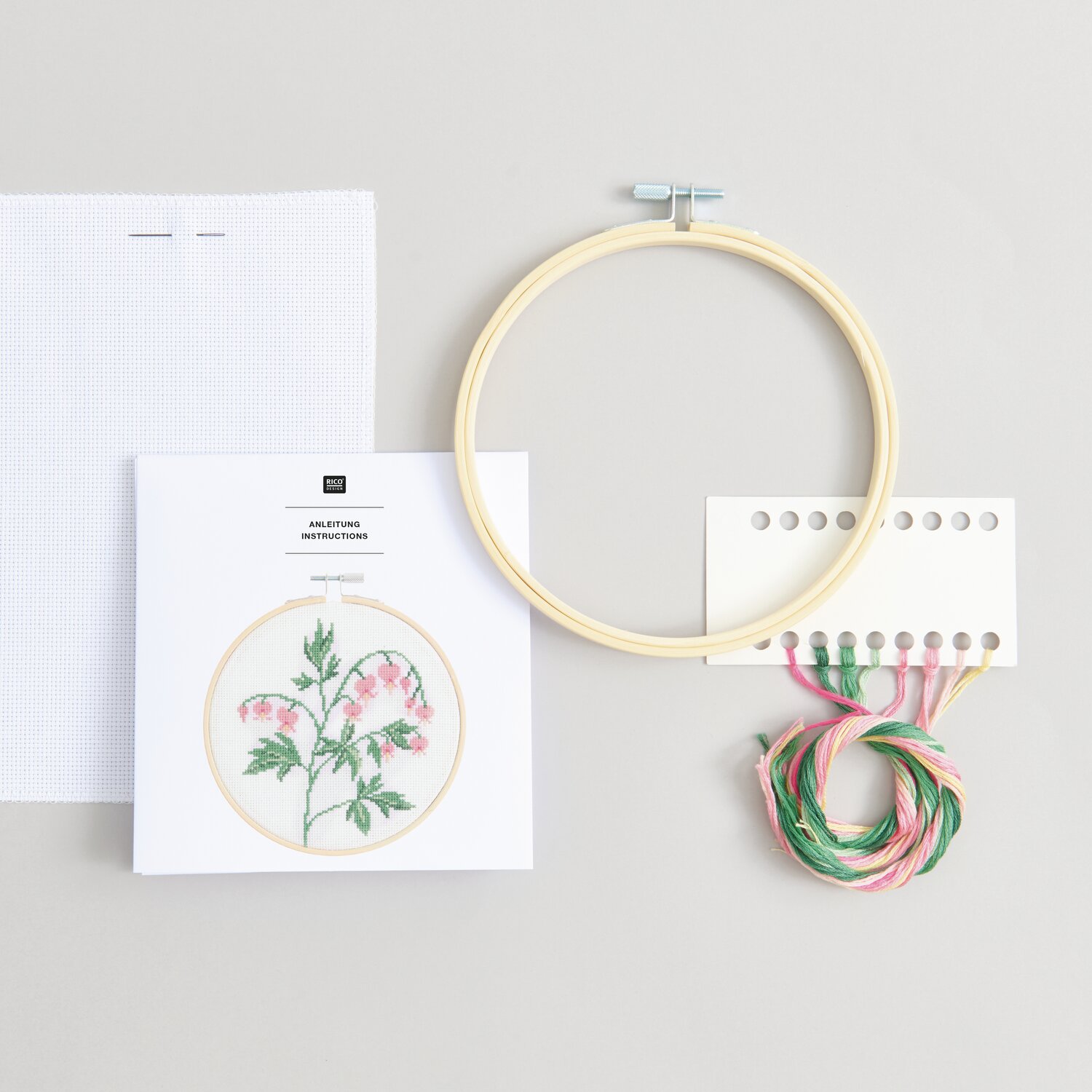 Stickpackung Gezählter Kreuzstich Tränendes Herz mit Stickrahmen, Garn und Anleitung von Rico Design auf grauem Hintergrund.