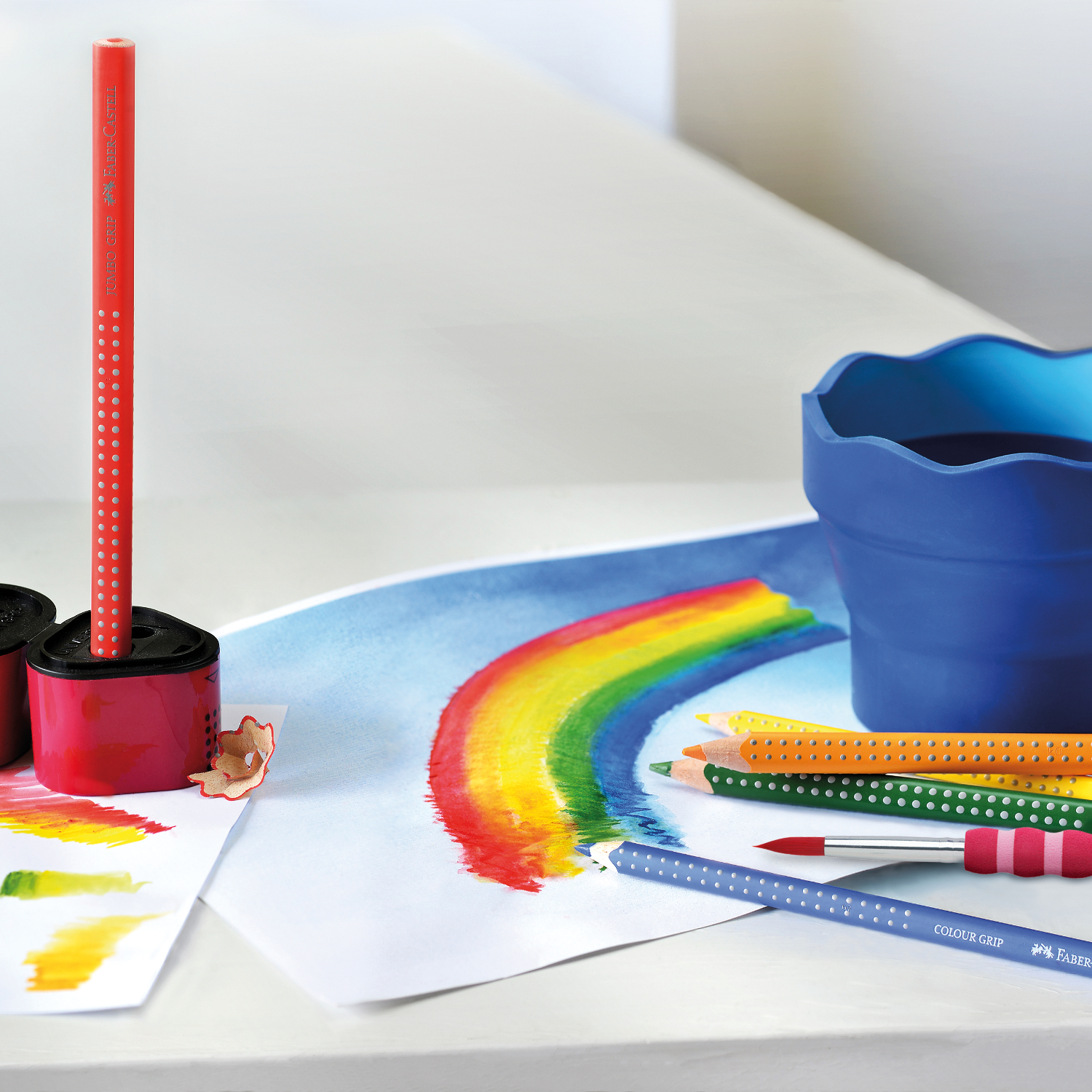 Jumbo Grip Farbstift, Faber Castell, in Helioblau rötlich, liegt neben einem gemalten Regenbogen auf weißem Papier.