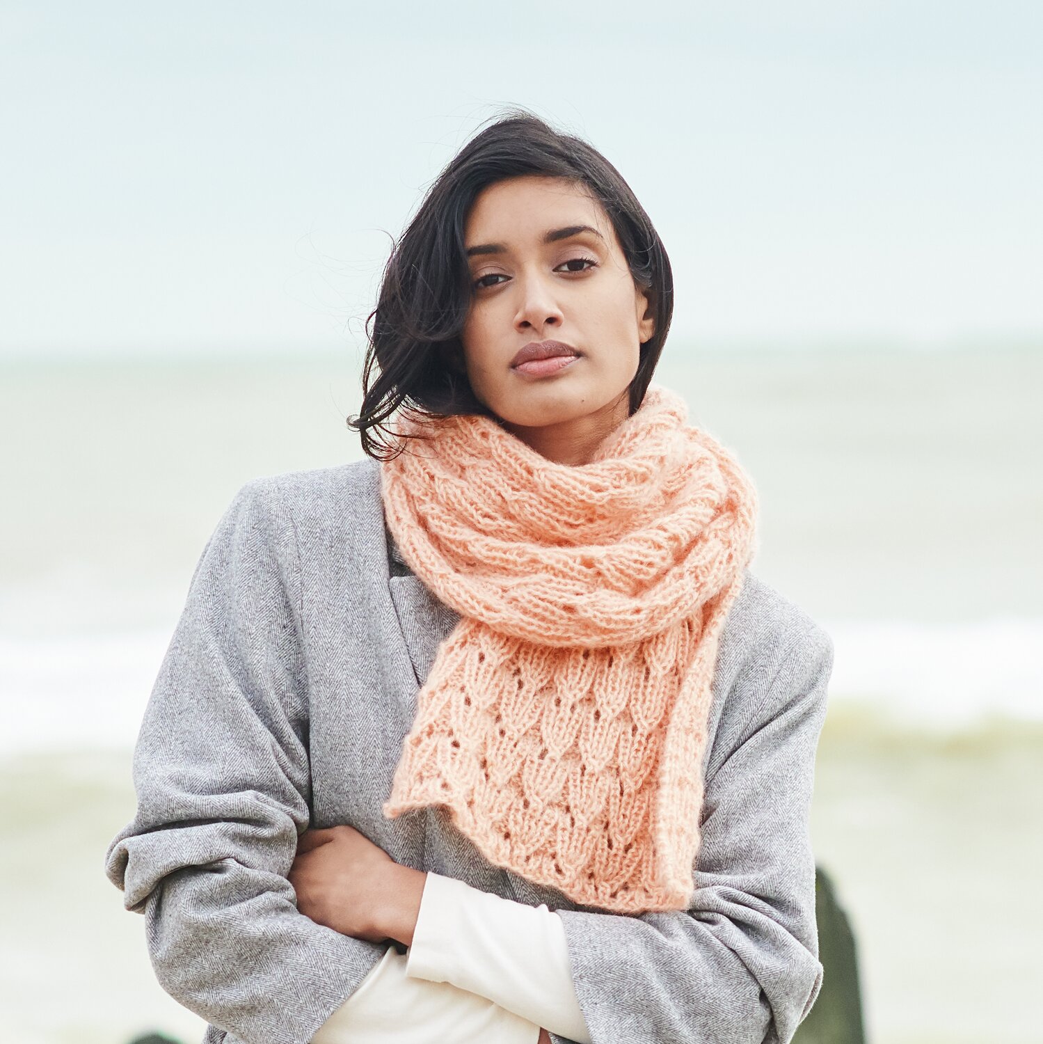 Strickset Schal Modell 39 aus Lovewool No. 21 von Rico Design Strickset Schal Modell 39 aus Lovewool No. 21: Frau trägt pfirsichfarbenen Schal am Strand, kombiniert mit grauem Mantel.