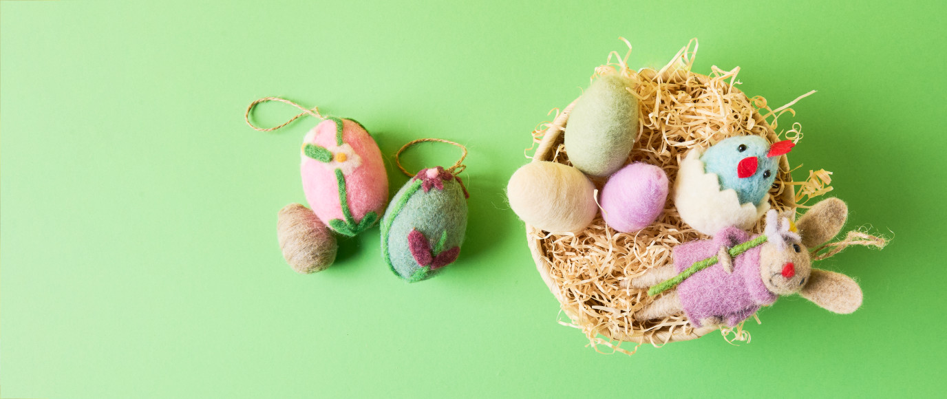 Ein Osterkorb gefüllt mit Ostergras, gefilzten Eiern und gefilzten Osterfiguren, daneben drei weitere gefilzte Eier. Flatlay von Produkten.