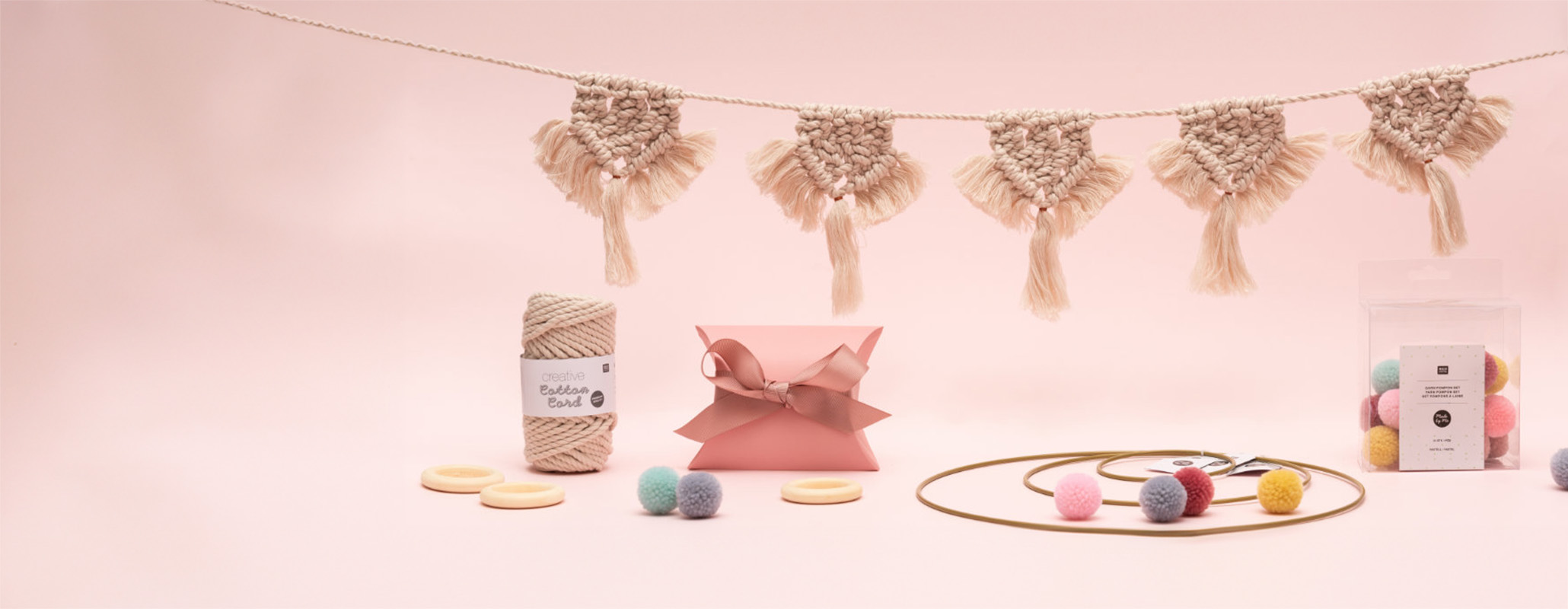 Beige Makramee-Girlande, Dekoringe und bunte Garn-Pompons. Hängende Makramee-Girlande mit Garn, eine Geschenkschachtel, verschiedene Dekoringe und Verpackung mit Garn-Pompons. Fotosetting rosa Wand.