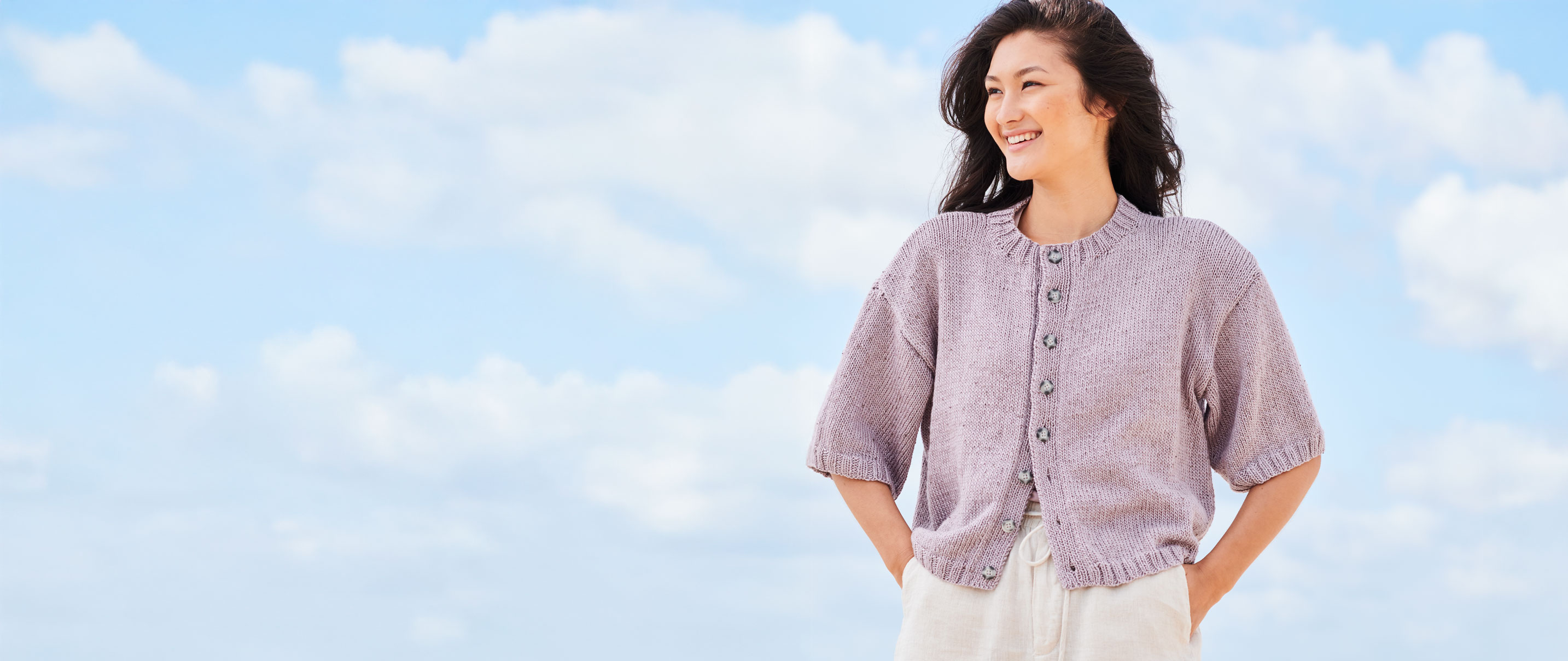 Frau mit fliederfarbener Strickjacke steht vor Wolken