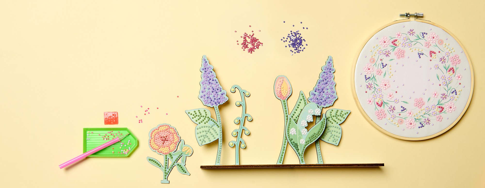 Ein florales Diamond-Painting auf einem Stickring und ein floraler Diamond-Painting-Dekoaufsteller, dazu das passende Zubehör. Flatlay von Produkten.