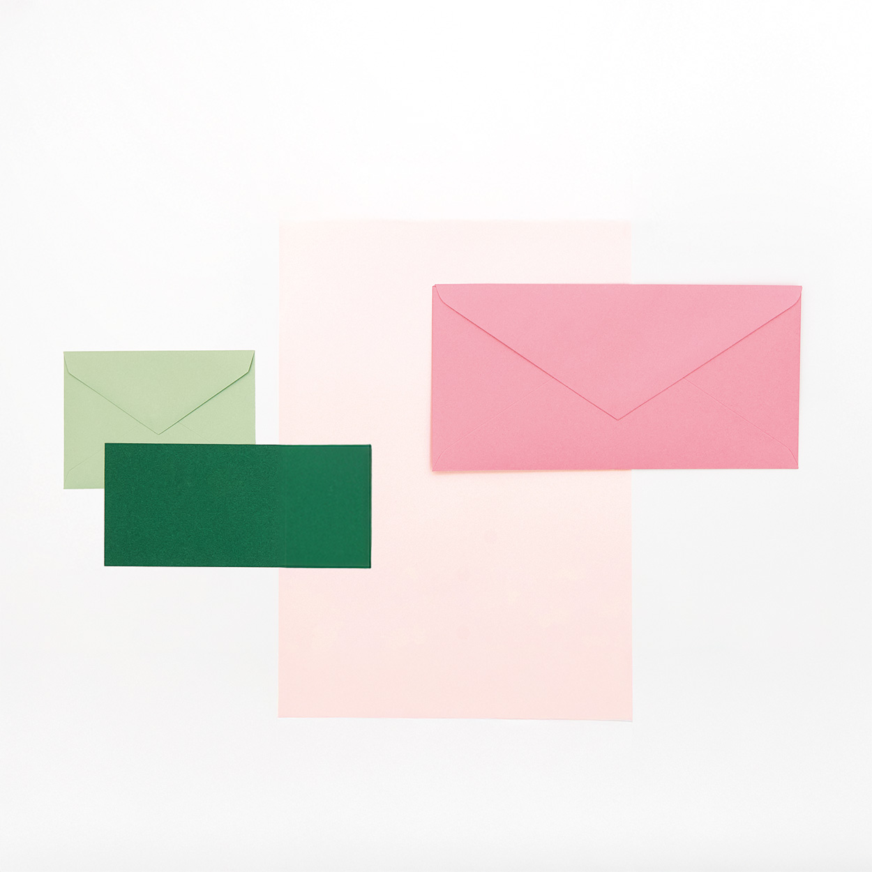 Ein Blatt rosa Papier darauf ei großer pinker Umschlag, sowie ein grüner Umschlag mit passender Karte. Flatlay von Produkten.