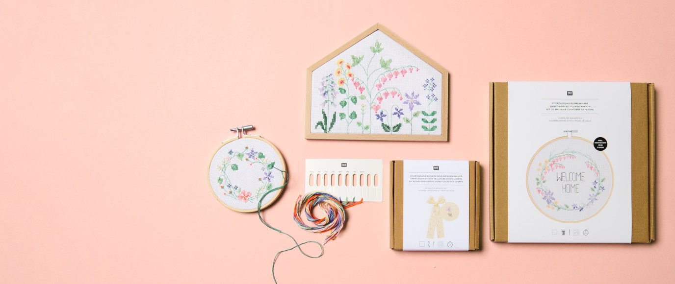Eine Stickpackung mit Stickring und Blumenkranz Motiv, sowie ein Stickrahmen in Hausform mit Blumenwiese Motiv. Flatlay von Stickpackungen.
