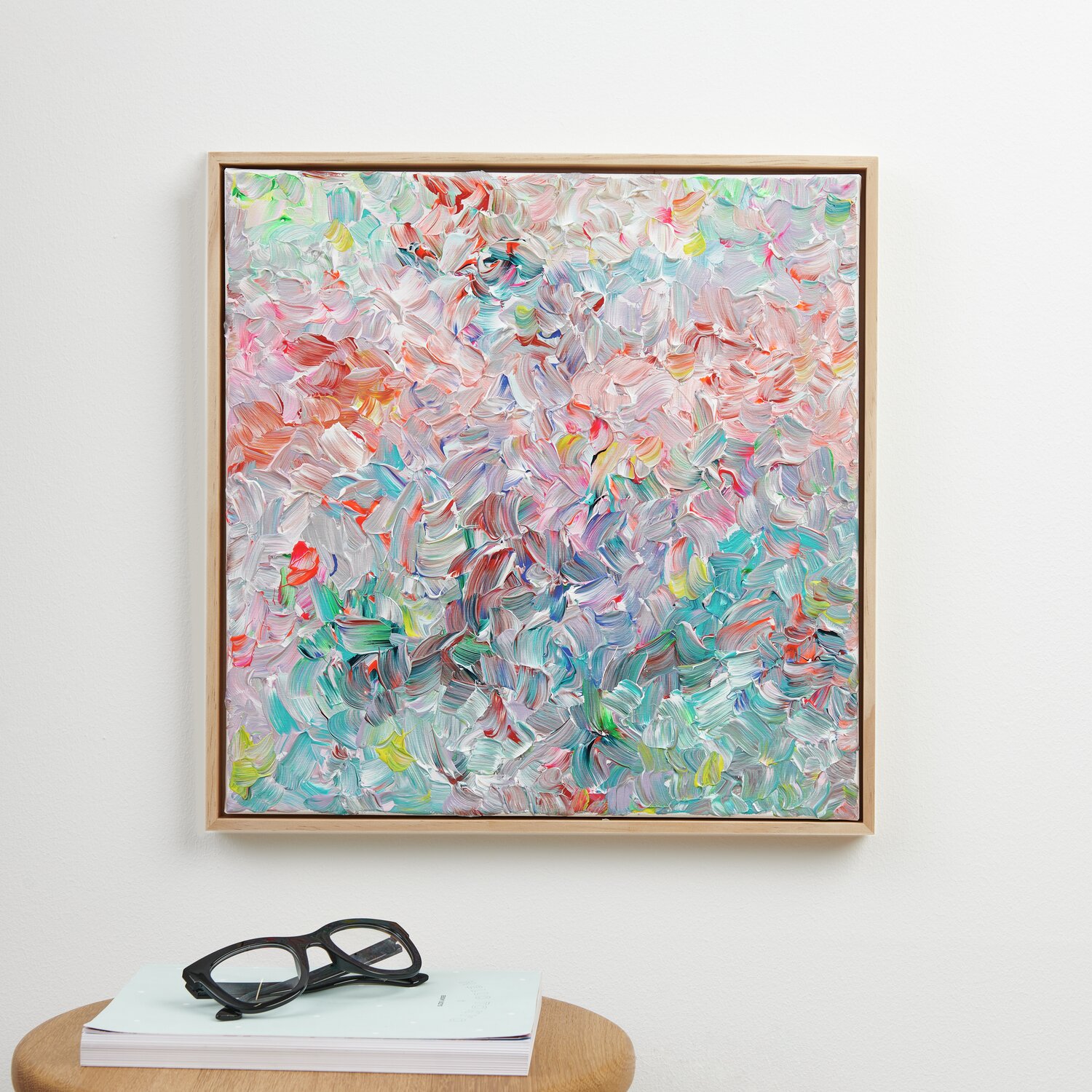 Abstraktes Gemälde in Pastelltönen, gerahmt an weißer Wand. Darunter Tisch mit Brille und Buch.