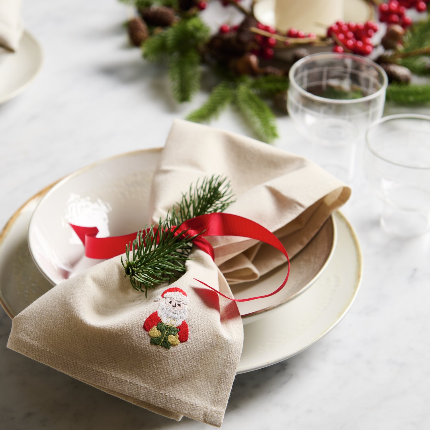 Stick & Stitch Stickpackung Weihnachtsmann zeigt ein besticktes Weihnachtsmotiv auf einem beigen Stoff, dekoriert mit Tannenzweig und rotem Band.