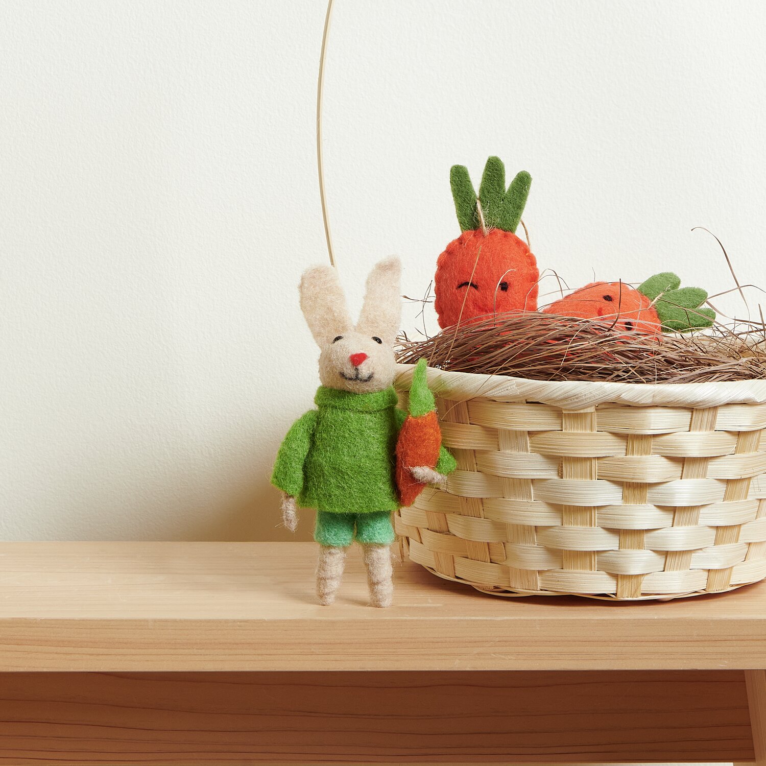 Aufhänger Filz-Hase mit Pulli und Möhre neben Bambus Korb mit Henkel Weiß, gefüllt mit Filz-Möhren, auf einem Holzregal.