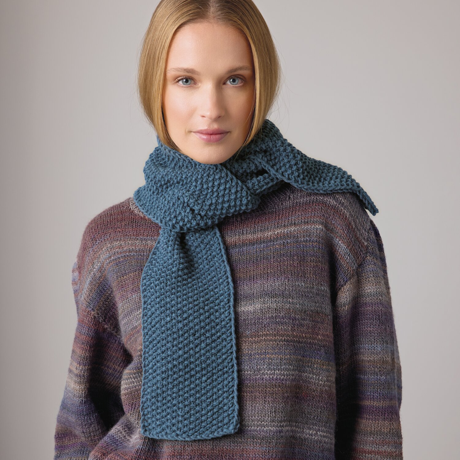 Strickset Mini-Schal Modell 14b aus Die Neue Masche No. 14 von idee. Creativmarkt Strickset Mini-Schal Modell 14b aus DNM 14: Frau trägt blauen Schal und mehrfarbigen Pullover.