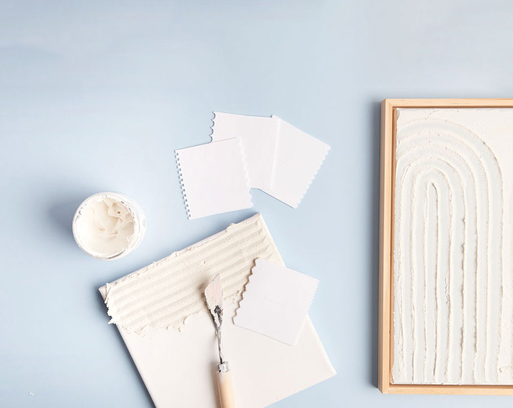Zwei Bilder mit Strukturpaste gestaltet, daneben verschiedene Effektspachtel und eine Dose mit Strukturpaste. Flatlay von Produkten.