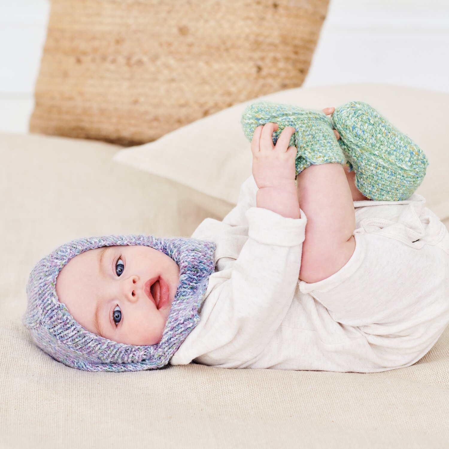 Häkelset Schuhe Modell 08b aus Baby 044 und Strickset Balaclava Modell 17a aus Baby 044, liegendes Baby mit weichen Stricksachen auf hellem Sofa