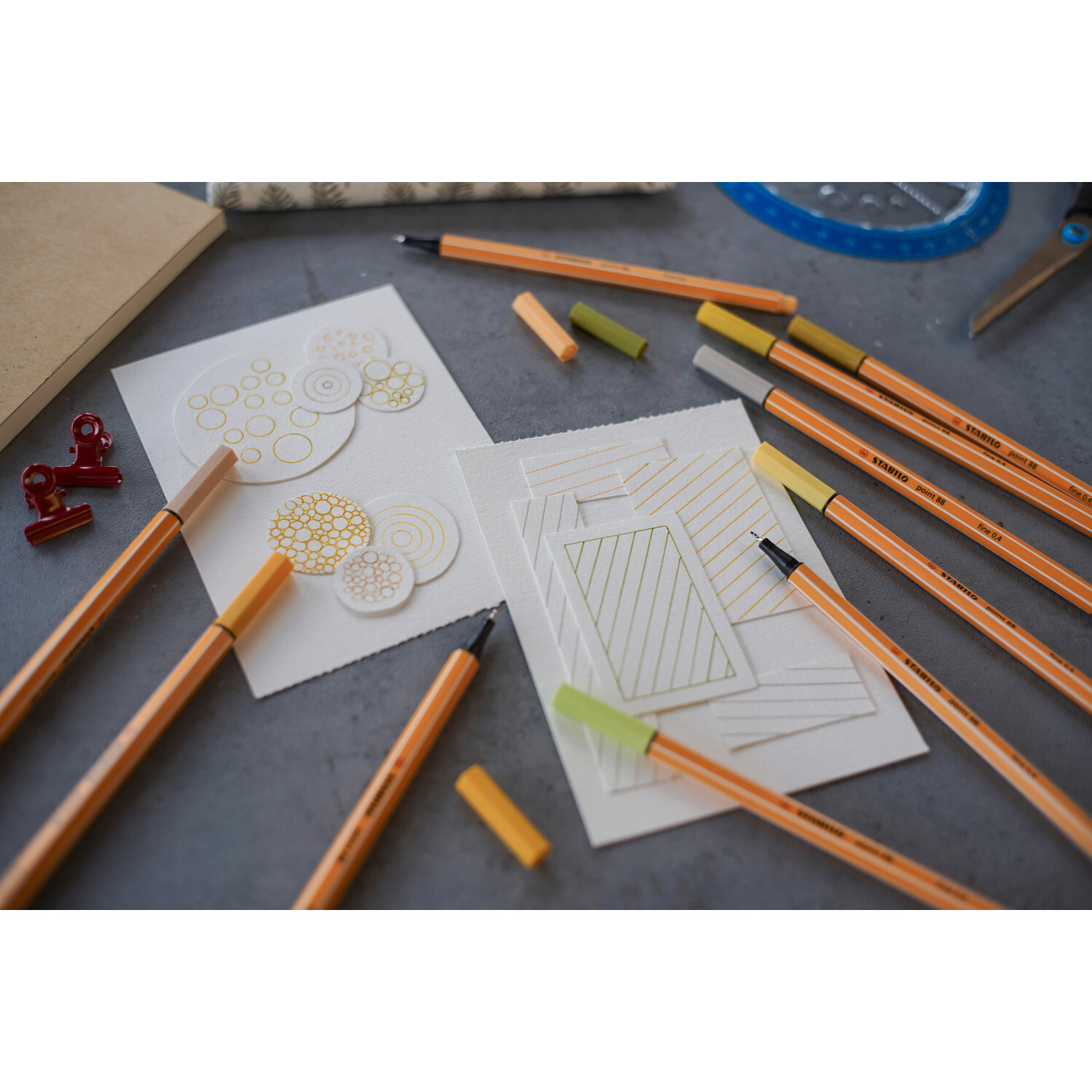 Point 88 Fineliner Set, 15-teilig, auf grauem Tisch mit Zeichnungen und orangefarbenen Stiften.