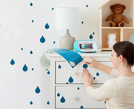 Frau beklebt eine Kommode mit Regentropfen, auf der Kommode steht ein Schneideplotter und Klebepapier.