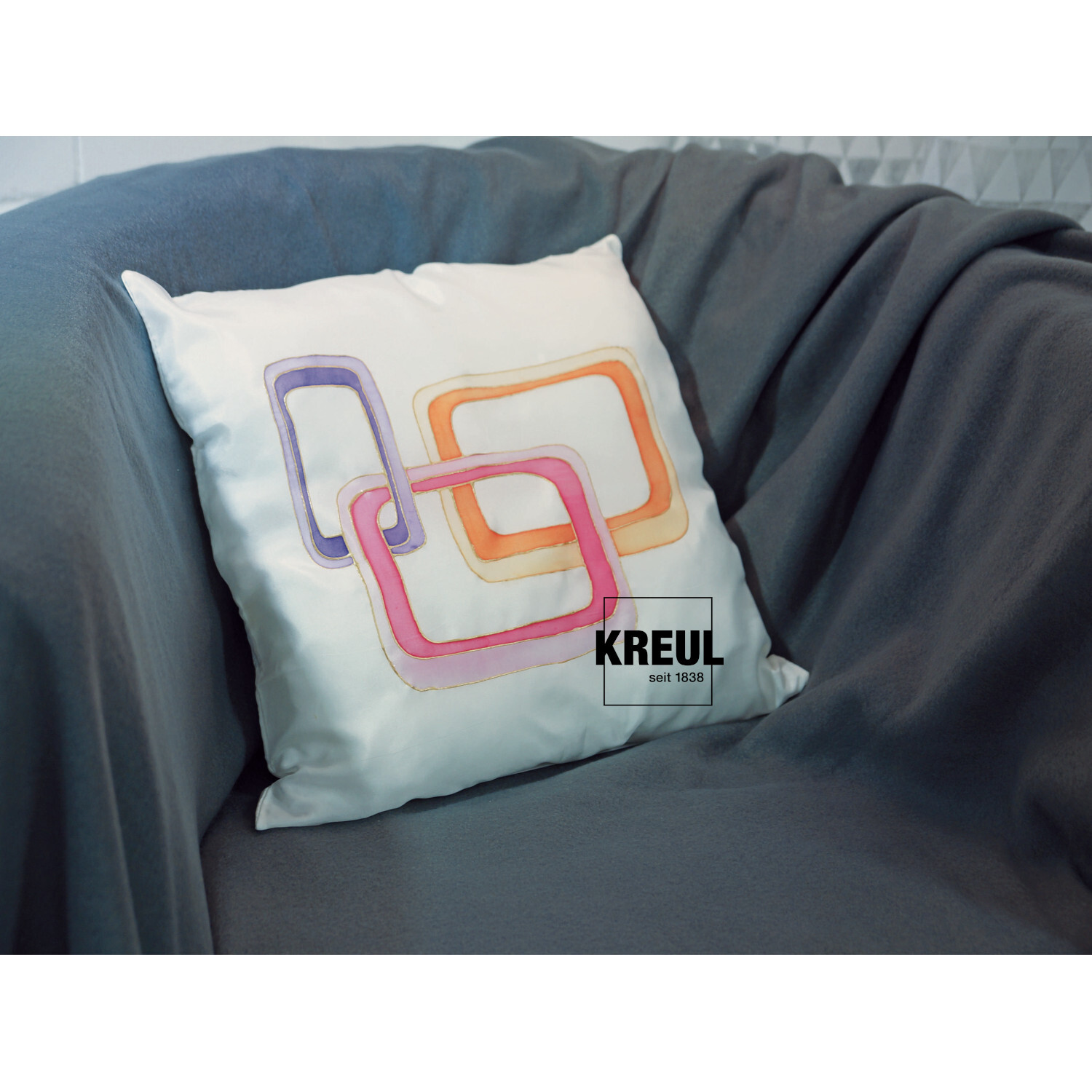 Javana Seidenmalfarben-Set Trendfarben: Weißes Kissen mit buntem, geometrischem Muster auf grauem Sofa. Marke: KREUL.