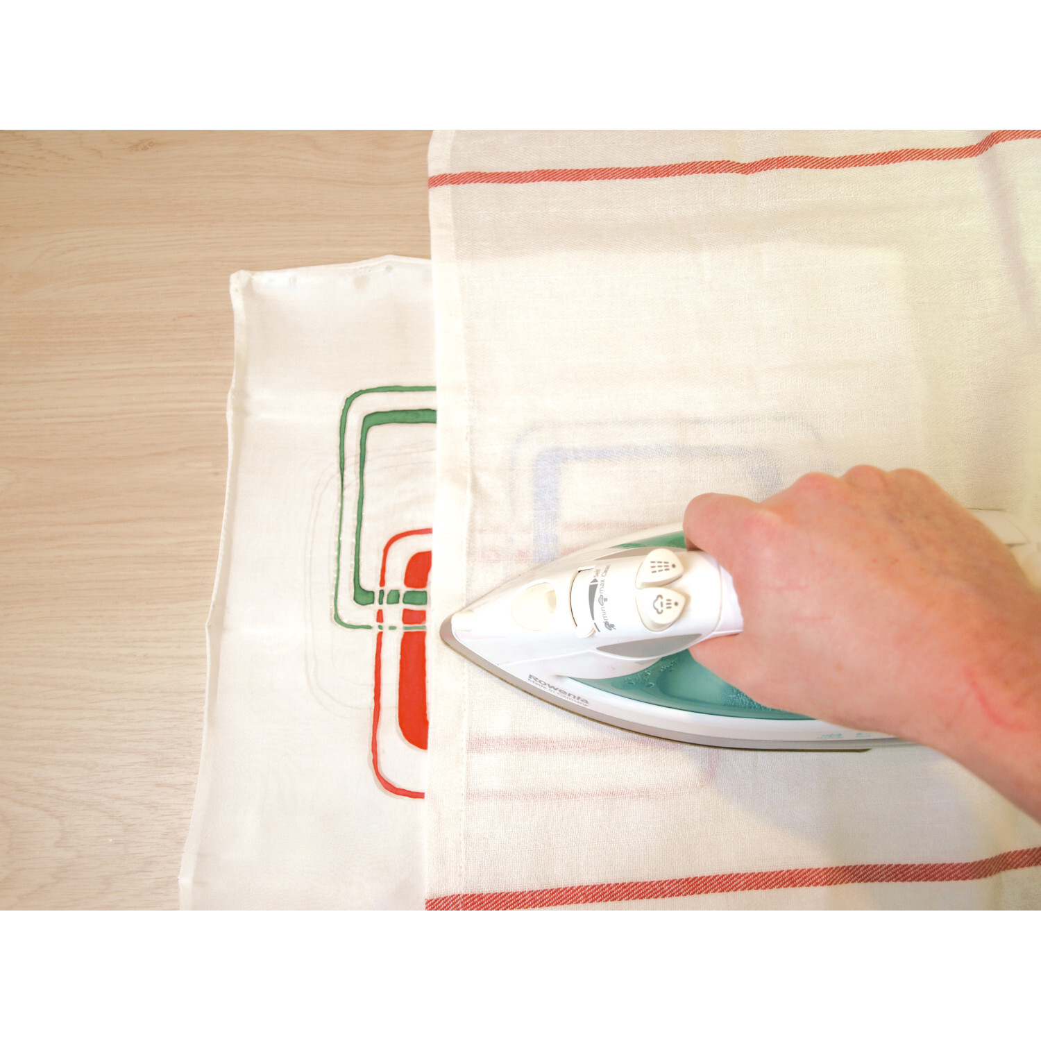 Javana Seidenmalfarben-Set Grundfarben: Eine Hand bügelt ein weißes Tuch mit buntem Muster auf Holz.