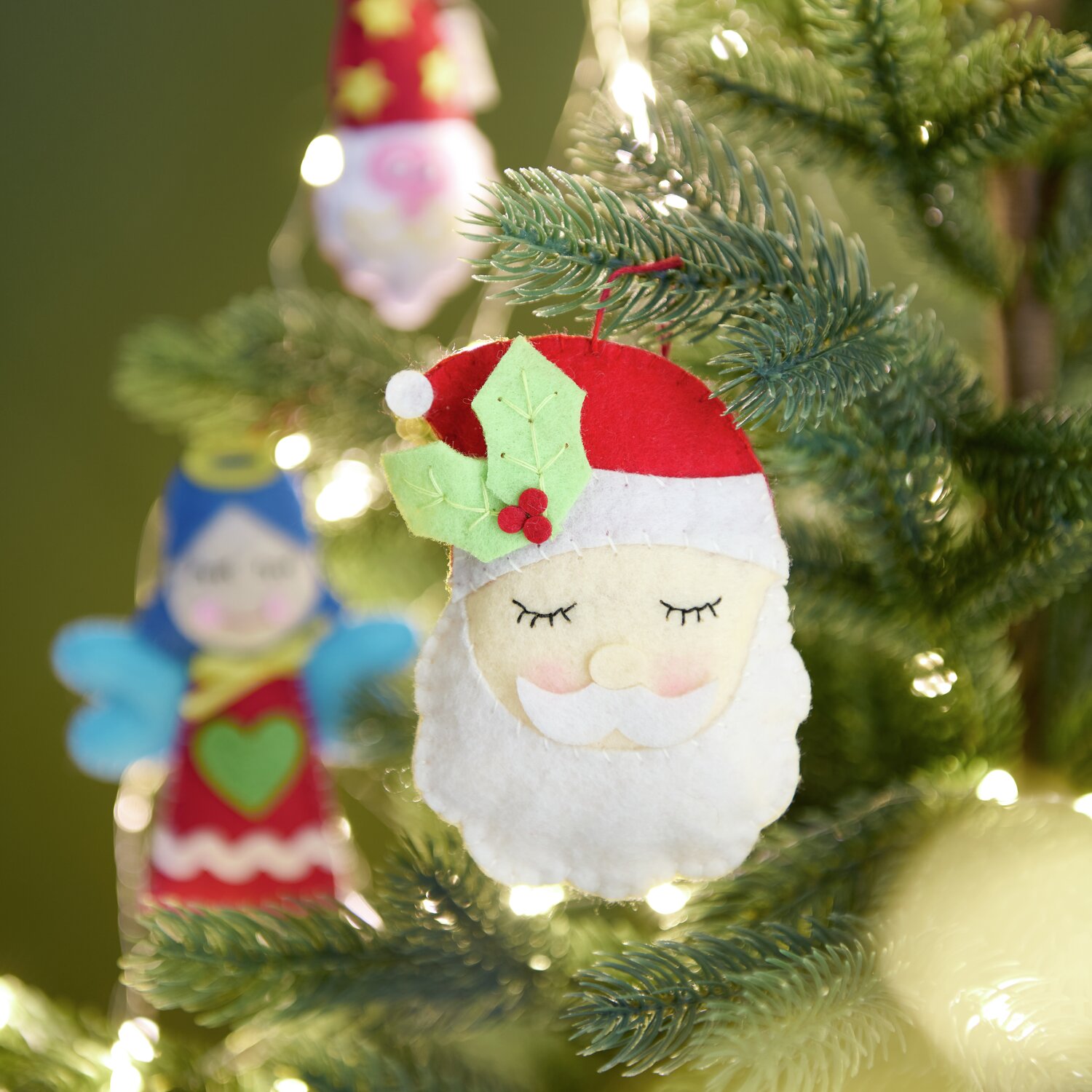 Aufhänger Filz-Santa mit Ilex Rot/Weiß: Weihnachtlicher Filzanhänger in Rot und Weiß, dekoriert mit Ilexblättern, am Weihnachtsbaum.