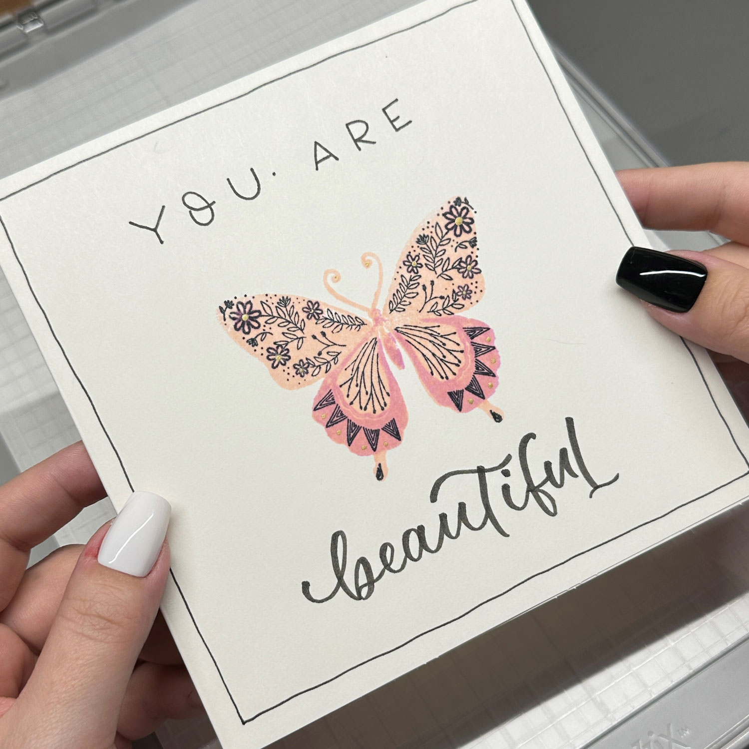 Eine Karte, mit einem rosa-orangenen gemalten Schmetterling und einem Schriftzug, wird von zwei Händen gehalten.