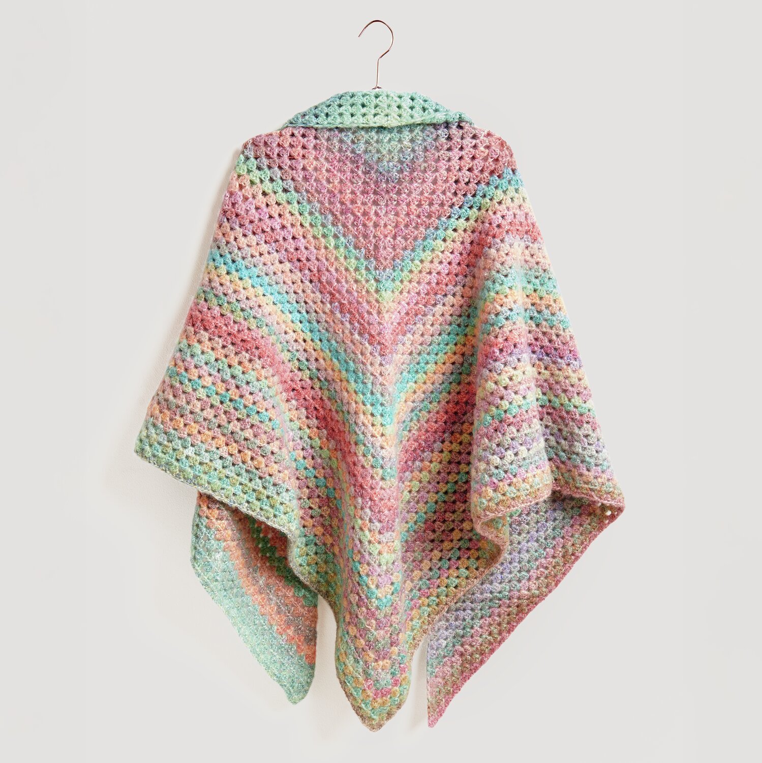 Die Neue Masche No. 13 Herbst von idee. Creativmarkt Häkelset Dreieckstuch Modell 11 zeigt ein buntes, handgefertigtes Tuch in Pastelltönen, aufgehängt an einem Haken.
