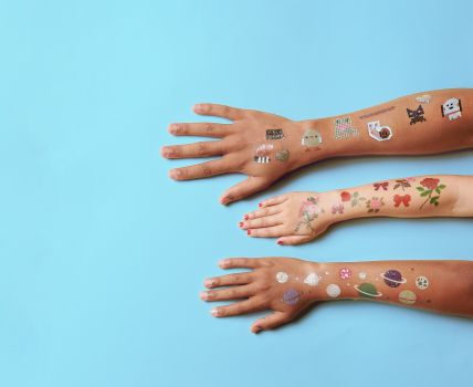 Drei ausgestreckte Arme auf hellblauem Hintergrund, verziert mit bunten temporären Tattoos von Rico Design mit verschiedenen Motiven wie Pixelgrafiken, Blumen und Planeten.
