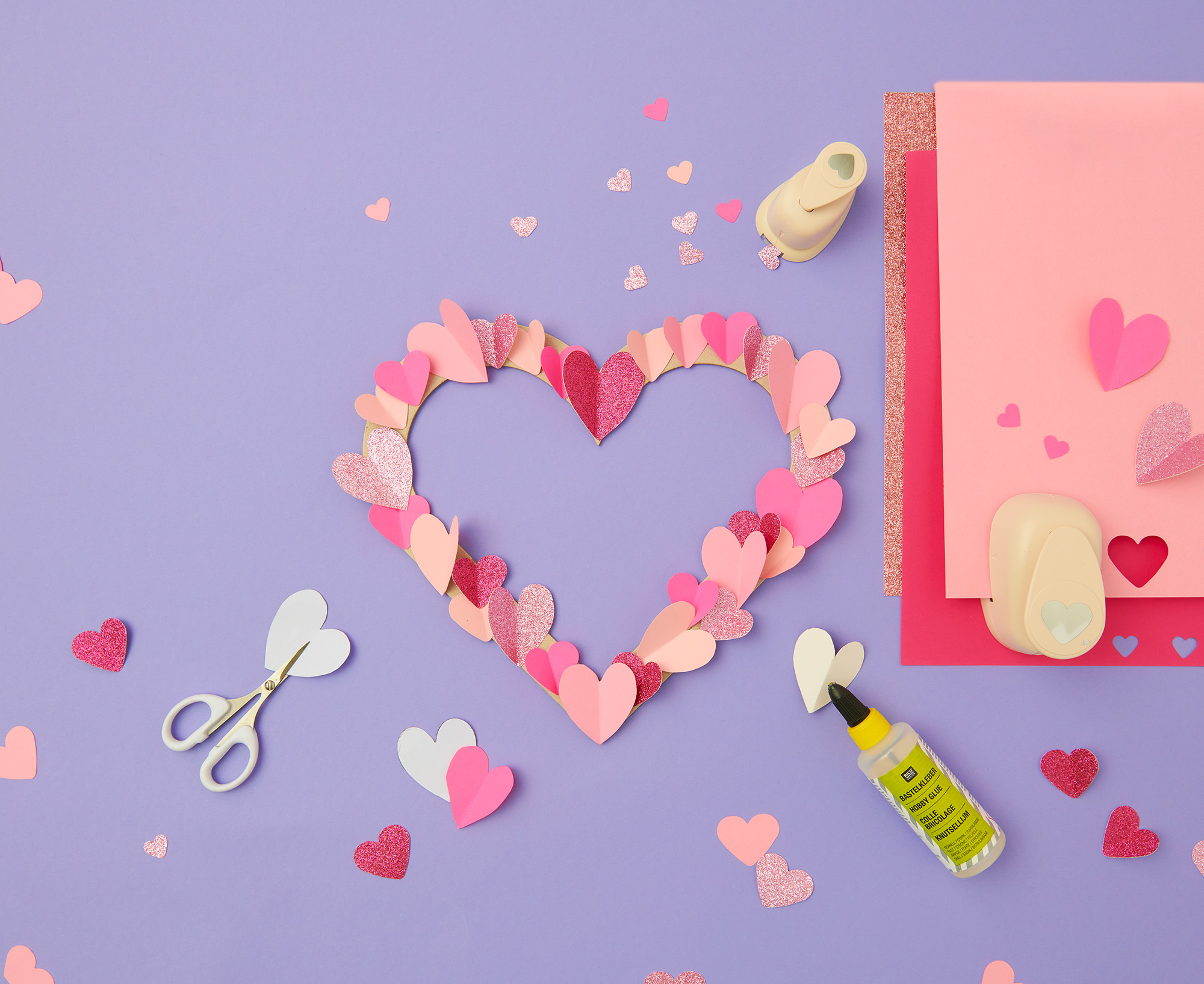 Herz aus mehreren Herzen in unterschiedlichen Farben und Größen, daneben Bastelpapier mit Herz-Stanzer und Kleber. Flatlay von Produkten.