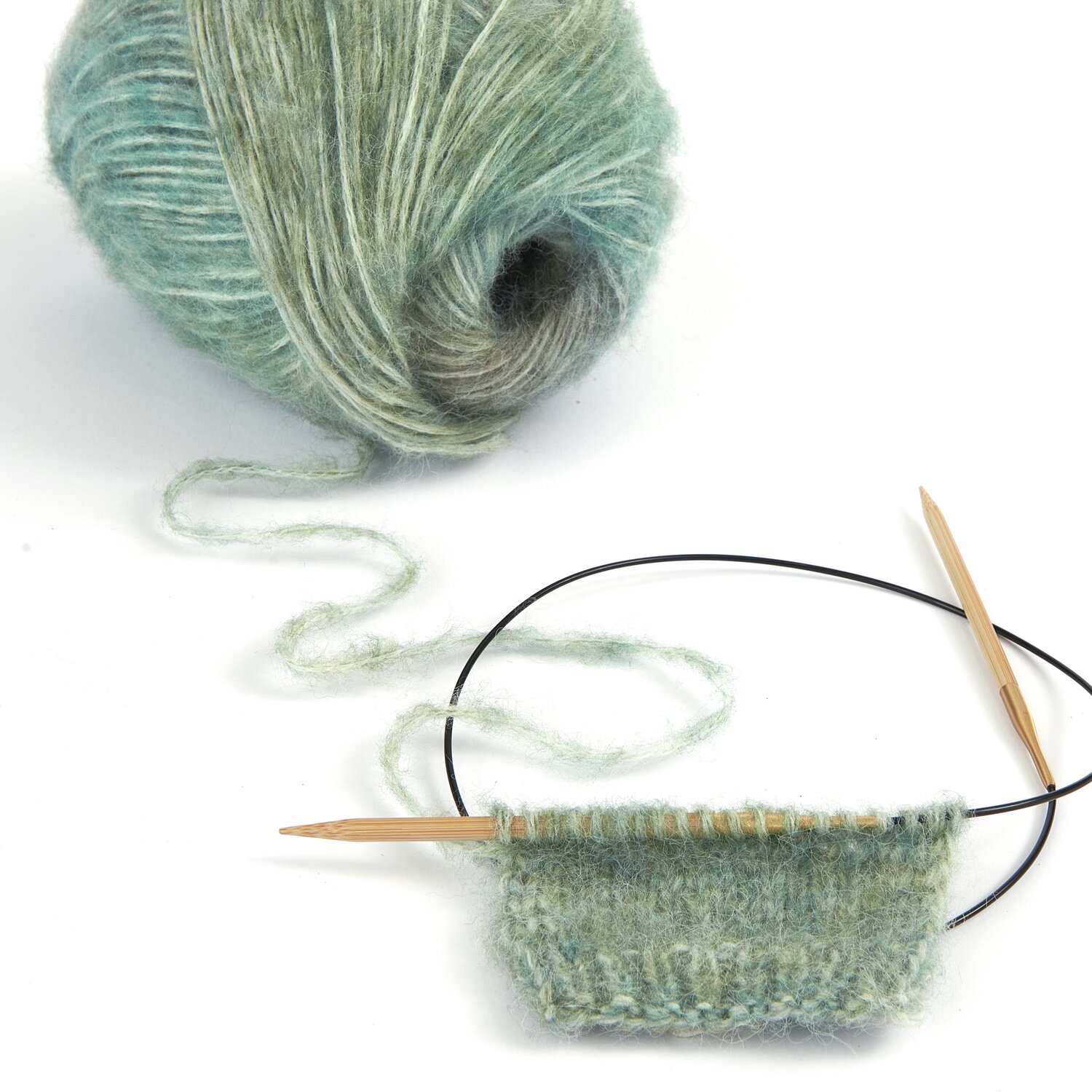 Wolle in sanftem Grün von [Marke], ideal für Strickprojekte. Gezeigt wird ein begonnenes Strickstück mit Rundstricknadeln. Perfekt für kreative Handarbeiten.