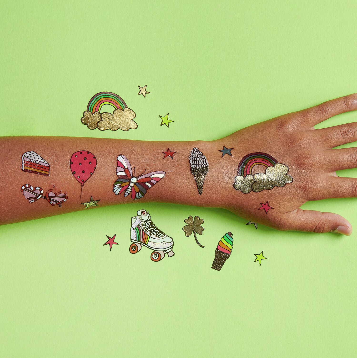 Tattoos Rainbow Fun: Bunte temporäre Tattoos auf einem Arm, darunter Regenbogen, Schmetterling und Eis. 19-teiliges Set auf grünem Hintergrund.
