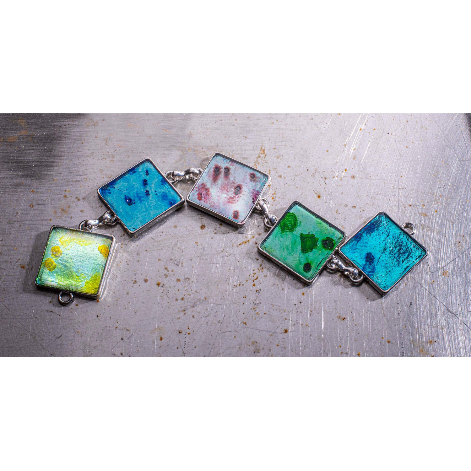 Crystal-Gießharz: Fünf bunte, quadratische Anhänger aus Harz auf Metalloberfläche, in Blau, Rot und Grün.