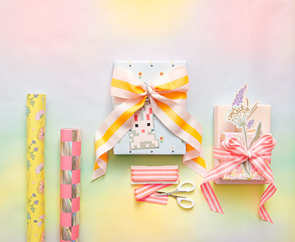 Zwei verpackte Geschenke mit Geschenkanhänger und Schleife, daneben zwei Geschenkpapierrollen, Geschenkband und eine Schere. Flatlay von Produkten.