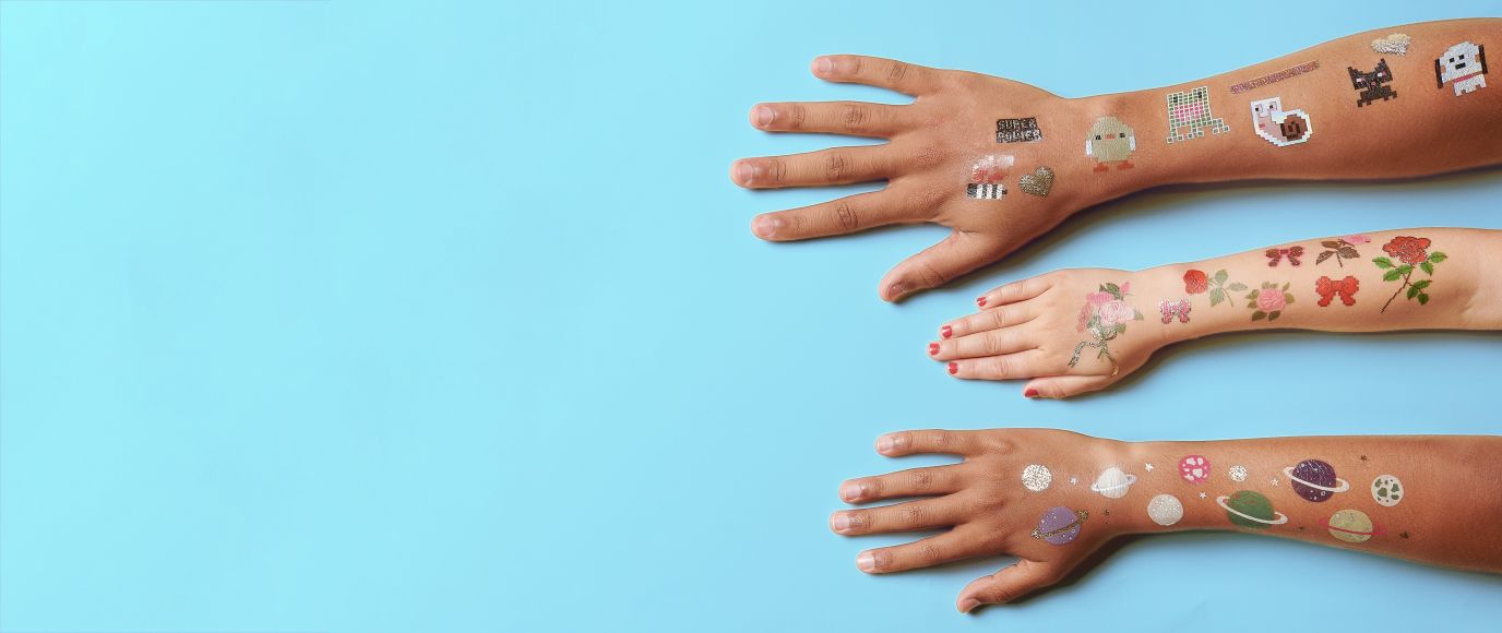 Drei ausgestreckte Arme auf hellblauem Hintergrund, verziert mit bunten temporären Tattoos von Rico Design mit verschiedenen Motiven wie Pixelgrafiken, Blumen und Planeten.