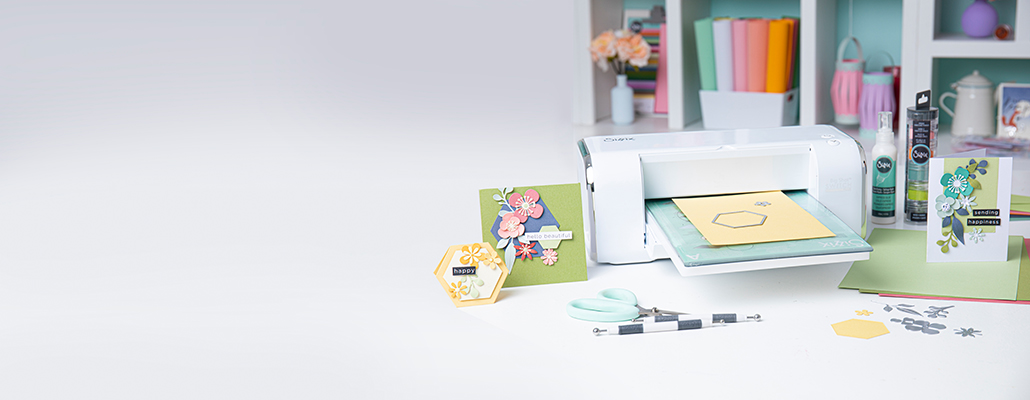 Stanzmaschine mit gelbem Papier und Framelit, sowie verschiedene Thinlits und Modellierzubehör, daneben gebastelte Karten mit Blumen-Design aufgestellt.