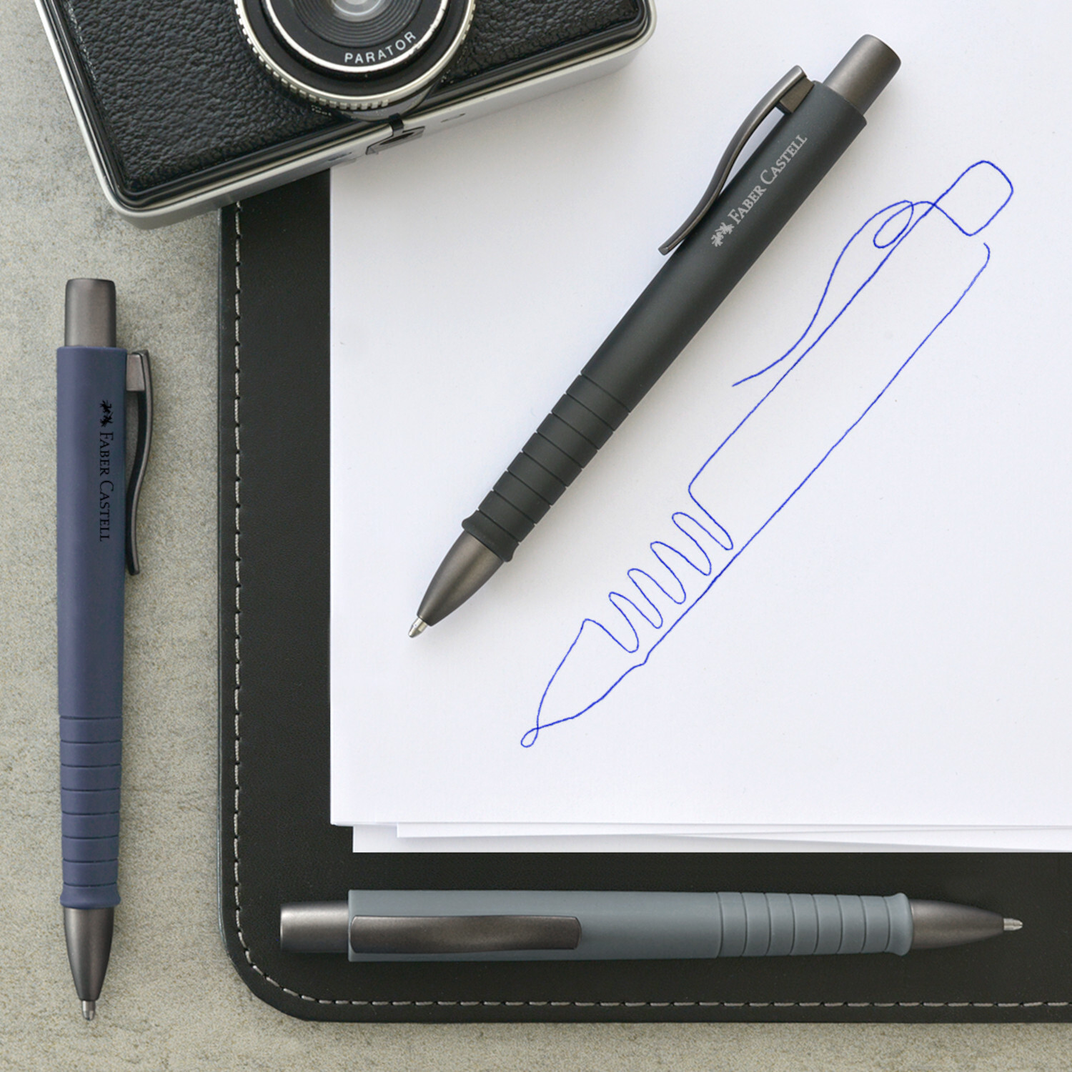 Kugelschreiber Poly Ball Urban XB Farbe Stone Grey von Faber-Castell Kugelschreiber Poly Ball Urban XB von Faber Castell in Blau und Schwarz auf Notizblock neben Kamera.