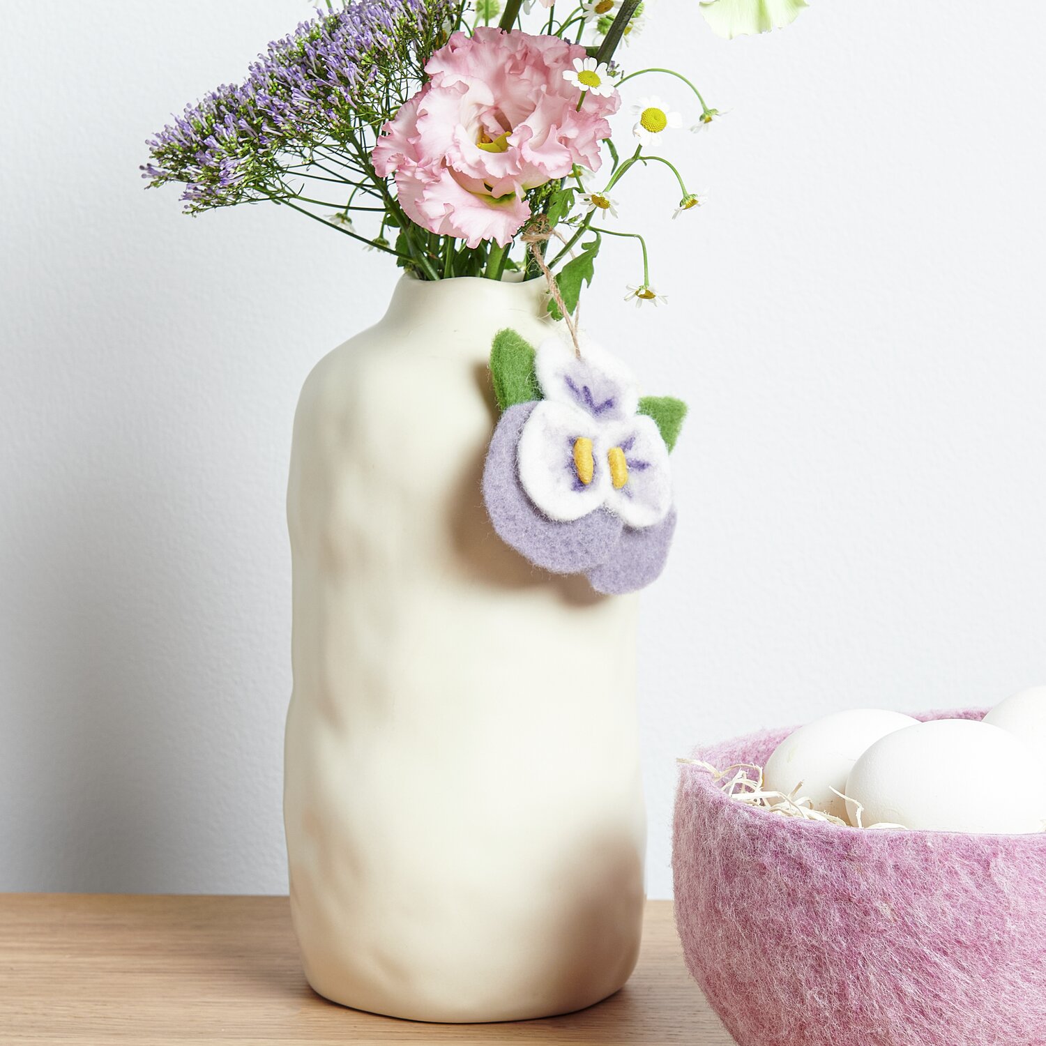 Aufhänger Filz-Blüte Stiefmütterchen an cremefarbener Vase mit Blumen, daneben rosa Filz-Körbchen mit Eiern.