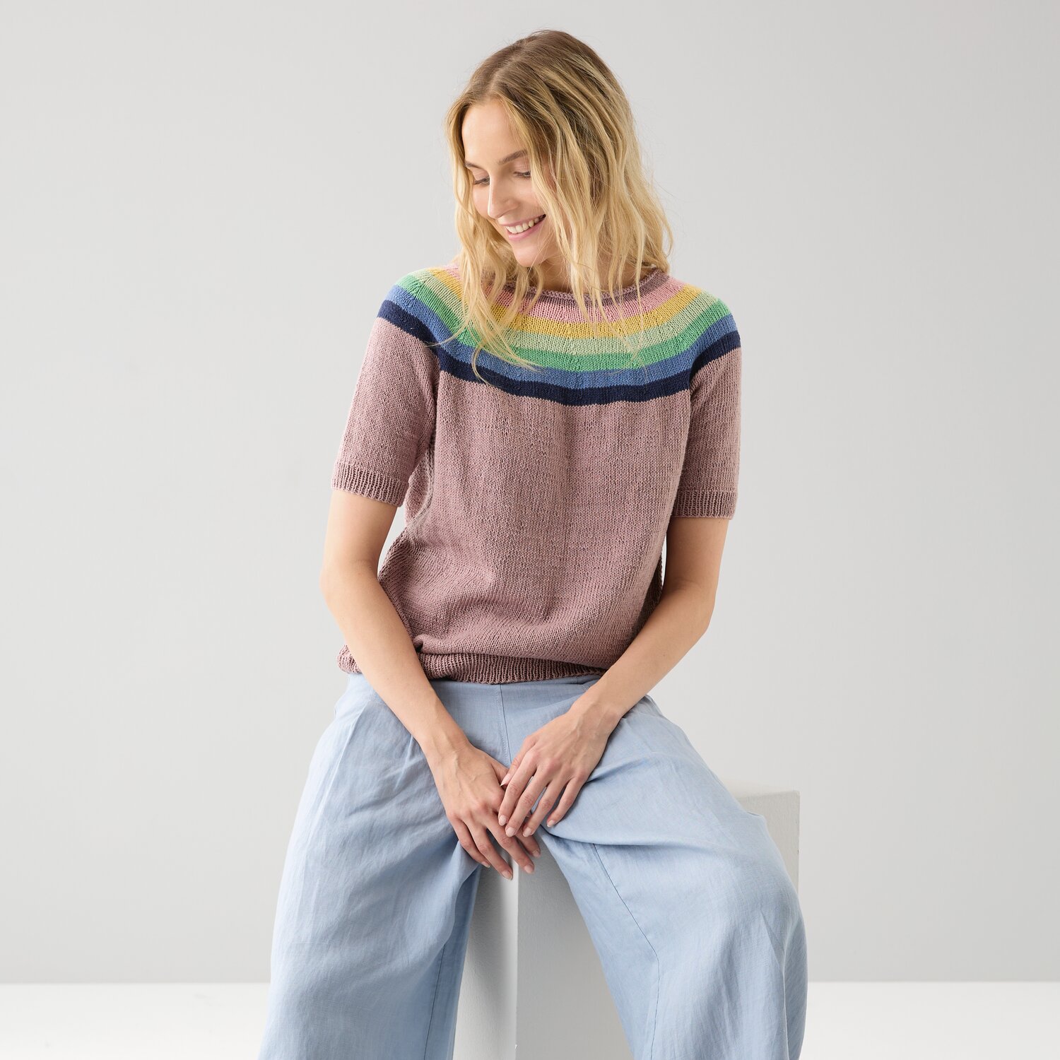Strickset Top-Down-Shirt Modell 22 A: Frau trägt ein buntes Shirt mit kurzen Ärmeln, sitzt lächelnd auf einem weißen Würfel.