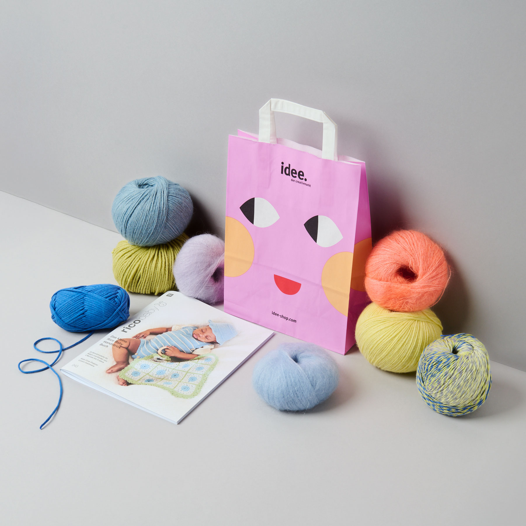 Eine Papiertüte daneben Garne in verschiedenen Farben und eine Zeitschrift. Fotosetting graue Ecke.