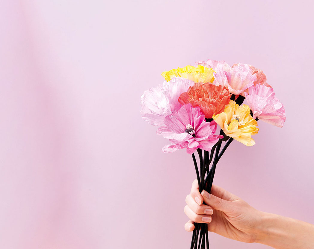 Eine Hand hält einen Blumenstrauß aus pinken, gelben und orangenen Papierblüten. Fotosetting pinker Hintergrund.