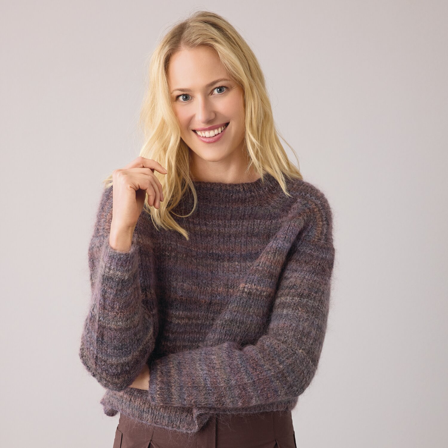 Strickset Pullover Modell 07 aus Die Neue Masche No. 14 von idee. Creativmarkt Strickset Pullover Modell 07 aus Die Neue Masche No. 14: Frau trägt lila gestrickten Pullover, lächelt und steht vor grauem Hintergrund.