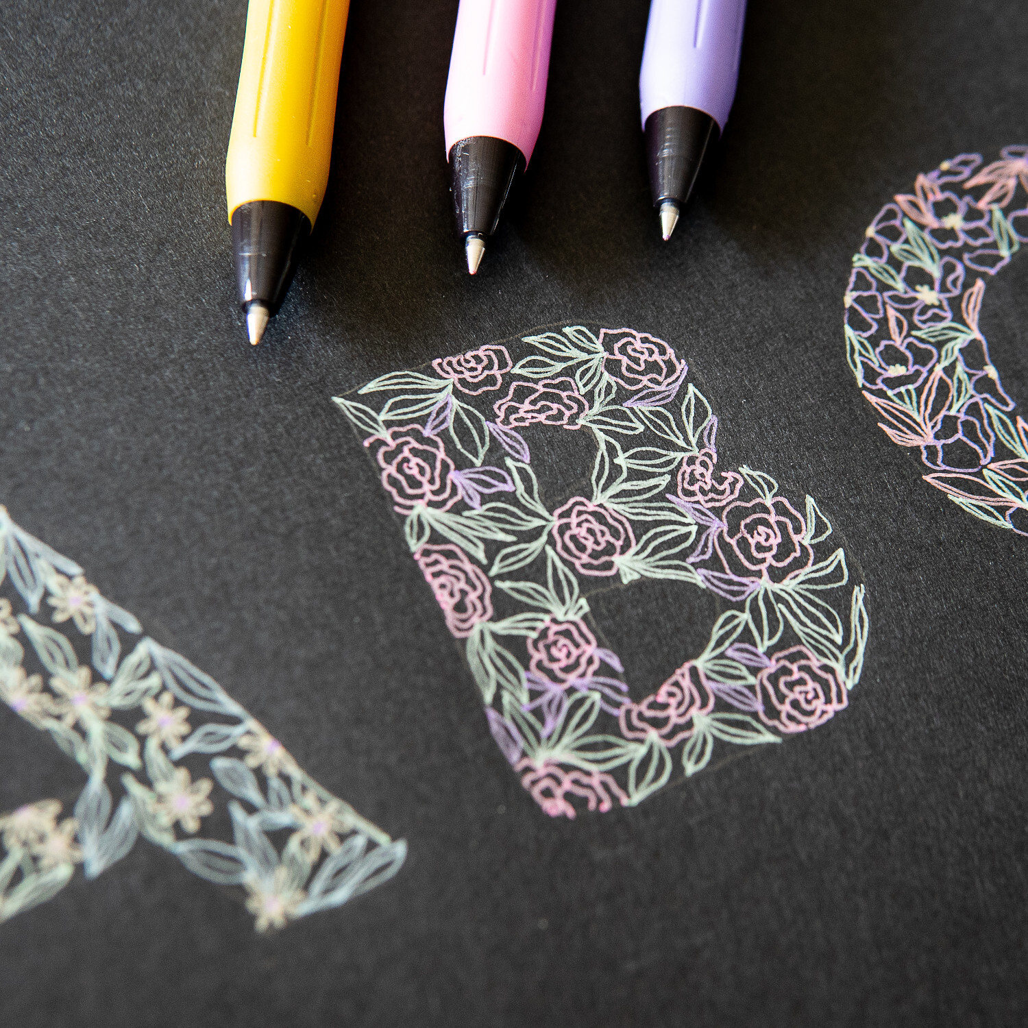 Sarasa Clip Set Decoshine: Drei bunte Stifte auf schwarzem Papier, verziert mit floralen Buchstaben.